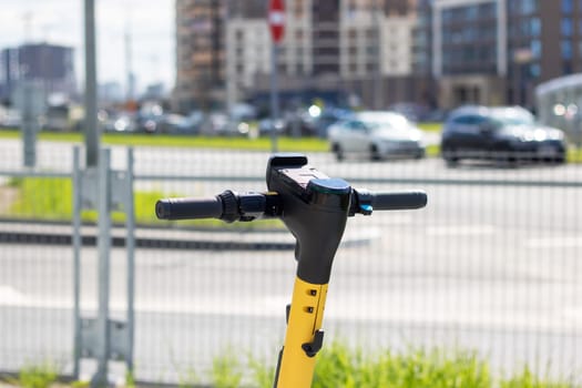 A black and yellow scooter with an electric blue bicycle handlebar is parked on the side of the asphalt road in the city, blending in with the metal car passing by. A vehicle for recreation