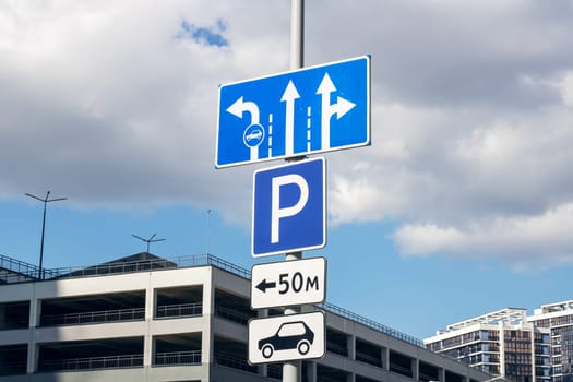 A blue parking sign is visible with arrows directing to the right. The sign is located in the city, near a building and a gas station. The sky is clear with cumulus clouds