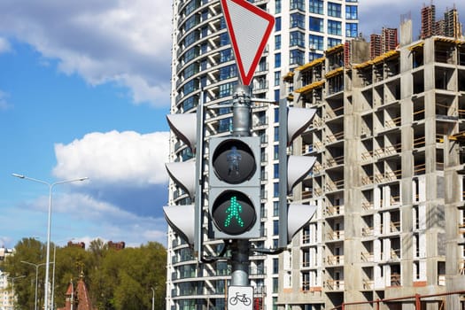 In front of a building under construction, a traffic light displays a green signal. The urban scene features a tower block, trees, and a cloudy sky