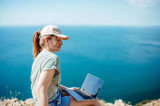 A woman is sitting on a rock by the ocean with a laptop in front of her. She is wearing a hat and has her hair in a ponytail. Concept of relaxation and leisure