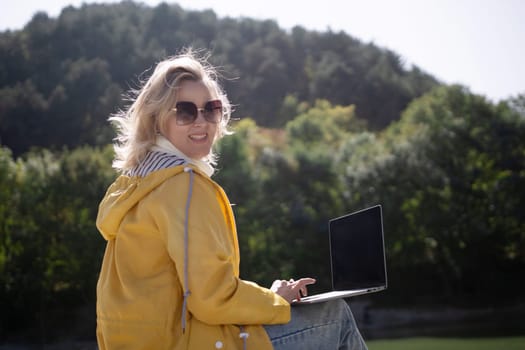 A woman in a yellow jacket is sitting on the grass with a laptop in front of her. She is smiling and she is enjoying her time outdoors