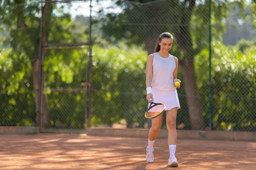 A woman is walking on a tennis court holding a tennis racket and a tennis ball. She is smiling and seems to be enjoying her time