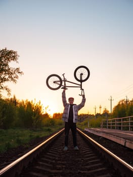 Silhouette of young BMX rider with his BMX cycle in hands above his head. BMX rider holds BMX bicycle with wheels up above head in sunset light, standing on railroad track. After BMX street riding