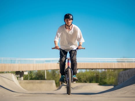 Young BMX bicycle rider having fun and rides in concrete skatepark outdoors. Skilled BMX freestyler in protective helmet posing in quarter pipe in ramp park. Cheerful boy rides on BMX, look at camera