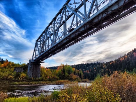 A steel bridge spans across a river in a colorful autumn forest under a clear blue sky