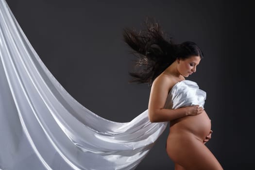 Naked pregnant woman posing with flying cloth, on gray background