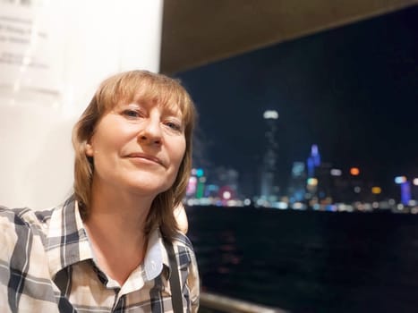 A middle-aged woman smiles for selfie with the Victoria Bay and Hong Kong skyline in the background at night