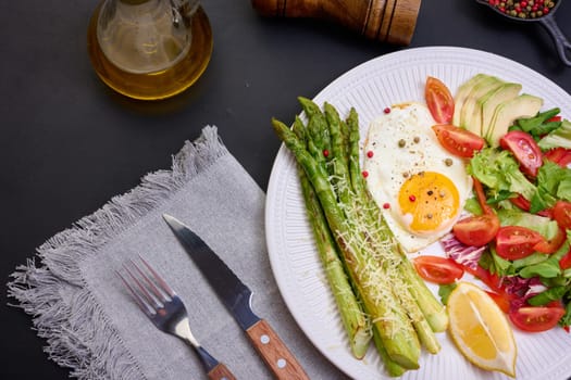 Round plate with cooked asparagus, fried egg, avocado and fresh vegetable salad on the table, top view