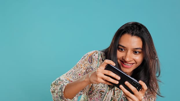 Indian woman playing videogames on cellphone in landscape mode, enjoying leisure time. Gamer enjoying game on mobile phone, having fun, isolated over studio background, camera B