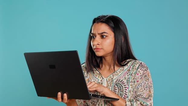 Indian woman typing on laptop keyboard and cursing, feeling angry at work. Person writing emails on notebook, complaining loudly, isolated over studio background, camera B