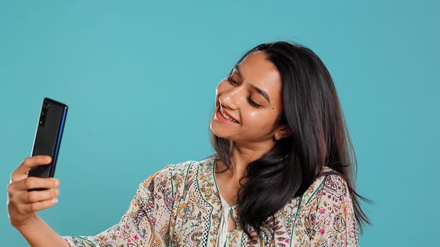 Jolly indian woman smiling for selfies, posting them on social media using cellphone. Joyous girl taking pictures using phone selfie camera, isolated over studio background, camera B