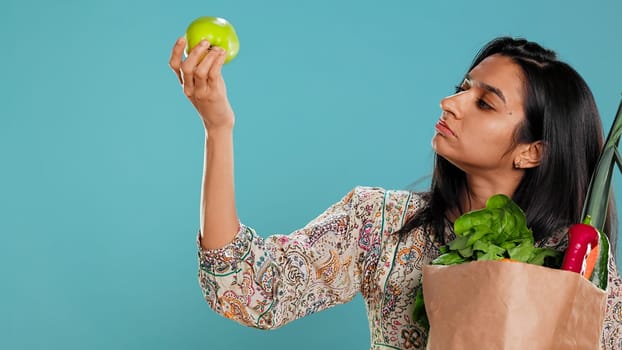 Indian woman with reusable paper bag in hands looking at apple, living healthy lifestyle. Ecology lover holding shopping bag with natural produce, studio background, camera B