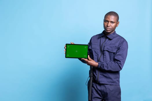 Security guard using a tablet to present an isolated greenscreen on camera, wearing his work uniform and smiling. Confident agent showing mockup screen on device in studio.