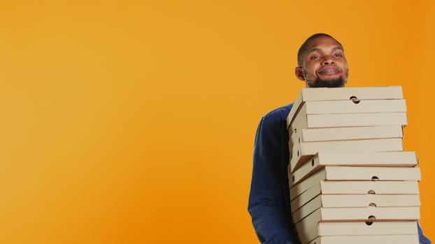 Confident male courier carrying big pile of pizza boxes to deliver fast food order to clients, takeaway service in studio. Young smiling deliveryman bringing takeout meal stack. Camera B.