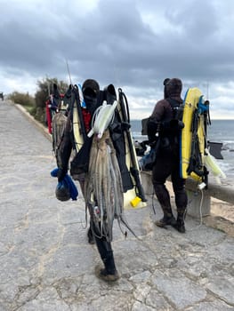 Back view of anonymous underwater fisherman in wetsuits and gears walking on footpath near sea under cloudy sky. Rear view of male friends with spearguns and catch leaving after hunting.