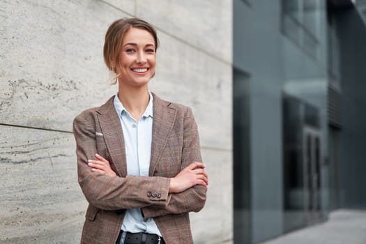 Portrait of smiling young businesswoman in formal standing arms crossed. Cheerful confident female entrepreneur looking at camera while standing outside modern office building.