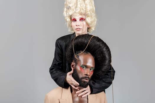 Confident gay man and woman, in vintage royal wigs and bold makeup, posing for studio portrait, looking directly at camera. Two LGBT friends in studio shot over light background.