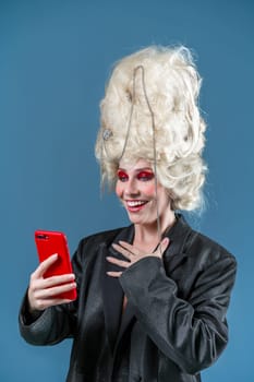 Smiling young woman in royal wig and red makeup using smartphone on blue background. Studio shot of happy funny girl in long jacket browsing phone surprise with good news.