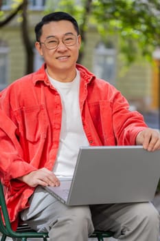 Asian middle-aged man freelancer working online distant job with laptop browsing website chatting sitting outdoors. Chinese guy on city street looking at notebook screen send messages watching movies