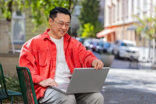 Asian middle-aged man freelancer working online distant job with laptop browsing website chatting sitting outdoors. Chinese guy on city street looking at notebook screen send messages watching movies