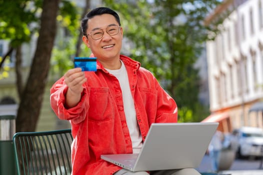 Asian middle-aged man using credit bank plastic card and laptop computer while transferring money purchases online shopping order food delivery on internet outdoors. Chinese guy on urban city street