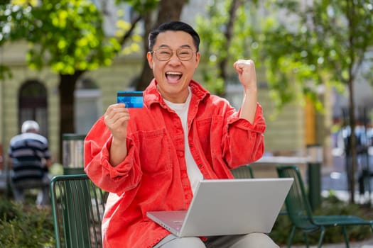 Asian middle-aged man using credit bank plastic card and laptop computer while transferring money purchases online shopping order food delivery on internet outdoors. Chinese guy on urban city street