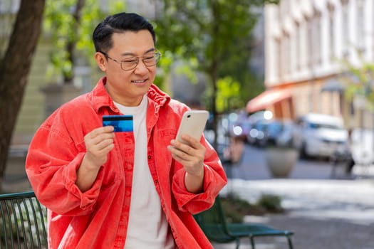 Asian middle-aged man using credit bank card smartphone while transferring money, purchases online shopping, order food delivery booking hotel room outdoors. Chinese guy on urban city street.