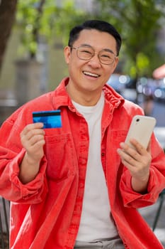 Asian middle-aged man using credit bank card smartphone while transferring money, purchases online shopping, order food delivery booking hotel room outdoors. Chinese guy on urban city street. Vertical