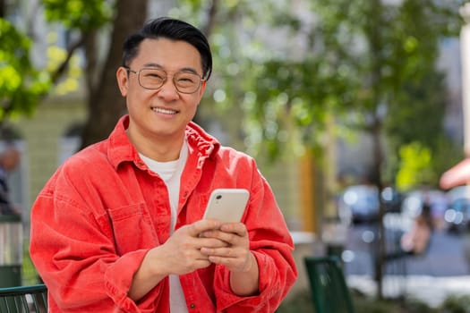 Asian middle-aged man using smartphone typing text messages in social media application online, surfing internet, relaxing, taking a break outdoors. Mature Chinese guy sitting on urban city street.