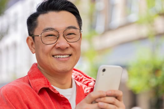 Close-up of Asian middle-aged man using smartphone typing text messages in social media application online, surfing internet, relaxing, taking a break outdoors. Chinese guy on city street. Horizontal