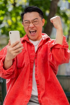 Asian middle-aged man use mobile smartphone celebrating win good message news, lottery casino jackpot victory, giveaway online outdoors. Chinese guy on urban city street. Town lifestyles. Vertical