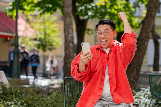 Asian middle-aged man use mobile smartphone celebrating win good message news, lottery casino jackpot victory, giveaway online outdoors. Chinese guy on urban city street. Town lifestyles. Horizontal