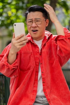 Sad Asian middle-aged man use smartphone typing browsing, fail loses lottery casino application, bad news results, fortune loss, outdoors. Chinese guy on urban city street. Town lifestyles. Vertical