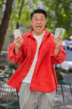 Happy rich Asian middle-aged man counting money dollar cash use smartphone calculator app plans to order gifts or food delivery online booking hotel room outdoors. Chinese guy on urban city street.