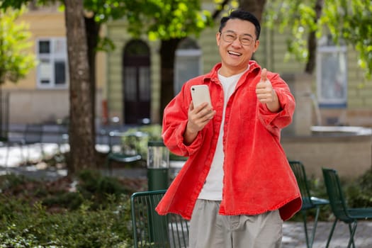 Like. Happy Asian middle-aged man raises thumbs up agrees with something gives positive reply recommends advertisement good idea feedback, celebrating success victory outdoors. Guy on city street.