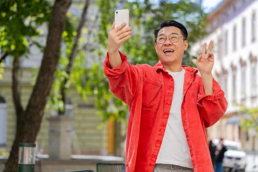 Asian middle-aged man blogger taking selfie on smartphone, communicating video call online with subscribers recording stories for social media vlog outdoors. Mature Chinese guy on urban city street.