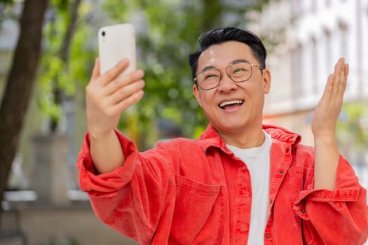 Asian middle-aged man blogger taking selfie on smartphone, communicating video call online with subscribers recording stories for social media vlog outdoors. Happy Chinese guy on urban city street.