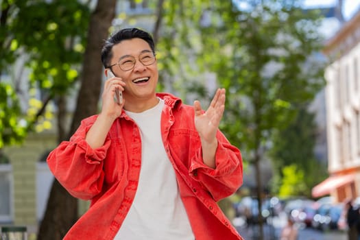 Happy smiling Asian middle-aged man having remote conversation communicate speaking by smartphone outdoors. Japanese guy talking on phone unexpected good news gossip on city street. Town lifestyles.