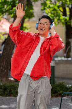 Happy overjoyed Asian middle-aged man dancing in wireless headphones listening favorite energetic music, celebrating victory win outdoors. Chinese guy on urban city street. Town lifestyles. Vertical