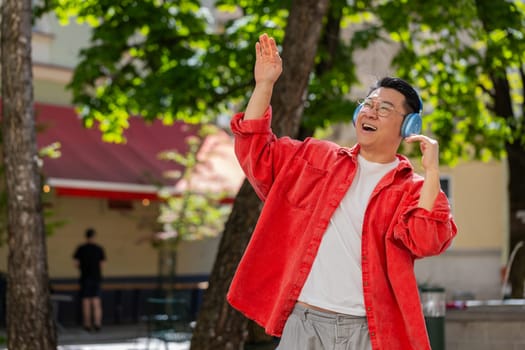 Happy overjoyed Asian middle-aged man dancing in wireless headphones listening favorite energetic music, celebrating victory win outdoors. Chinese guy on urban city street. Town lifestyles. Horizontal