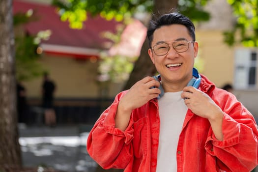 Portrait of happy smiling Asian middle-aged man listening music taking off headphones looking at camera relaxation feel satisfied good news outdoor. Chinese mature guy on urban city street. Horizontal