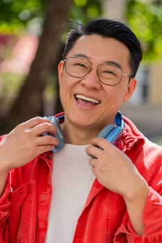 Portrait of happy smiling Asian middle-aged man listening music taking off headphones looking at camera, relaxation feel satisfied good news outdoors. Chinese mature guy on urban city street. Vertical