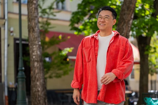 Asian middle-aged man tourist walking on urban city street outdoors in summer daytime. Chinese guy traveler smiling having positive good mood enjoying vacation holidays. Town lifestyles. Horizontal