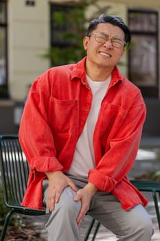 Sick Asian middle-aged man suffer from painful discomfort severe knee joint ache, leg muscles, could not walk outdoors. Chinese sad guy massaging knees illness cramps rheumatism on urban city street