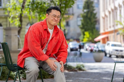 Sick Asian middle-aged man suffer from painful discomfort severe knee joint ache, leg muscles, could not walk outdoors. Chinese sad guy massaging knees illness cramps rheumatism on urban city street