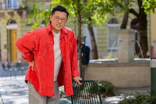 Asian middle-aged man suffering backache massaging sore place lower spine frowning from unbearable spasm torn muscles and tendons kidney stones pinched nerve outdoors. Chinese guy on urban city street