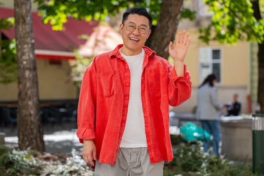 Hello. Asian middle-aged man smiling friendly at camera, waving hands gesturing hi, greeting or goodbye, welcoming with hospitable expression outdoors. Chinese guy on urban city street. Horizontal