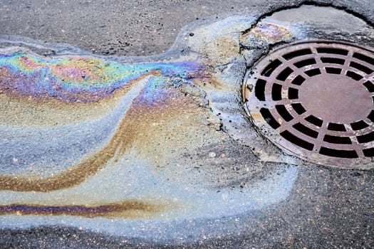 Fuel and lubricants from a vehicle mishap have created a hazardous puddle on the blacktop, flowing towards the nearby sewer grate.