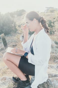 Digital nomad, woman in the hat, a business woman with a laptop sits on the rocks by the sea during sunset, makes a business transaction online from a distance. Freelance, remote work on vacation.