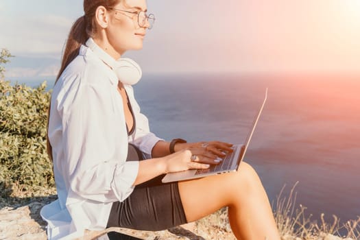 Digital nomad, woman in the hat, a business woman with a laptop sits on the rocks by the sea during sunset, makes a business transaction online from a distance. Freelance, remote work on vacation.